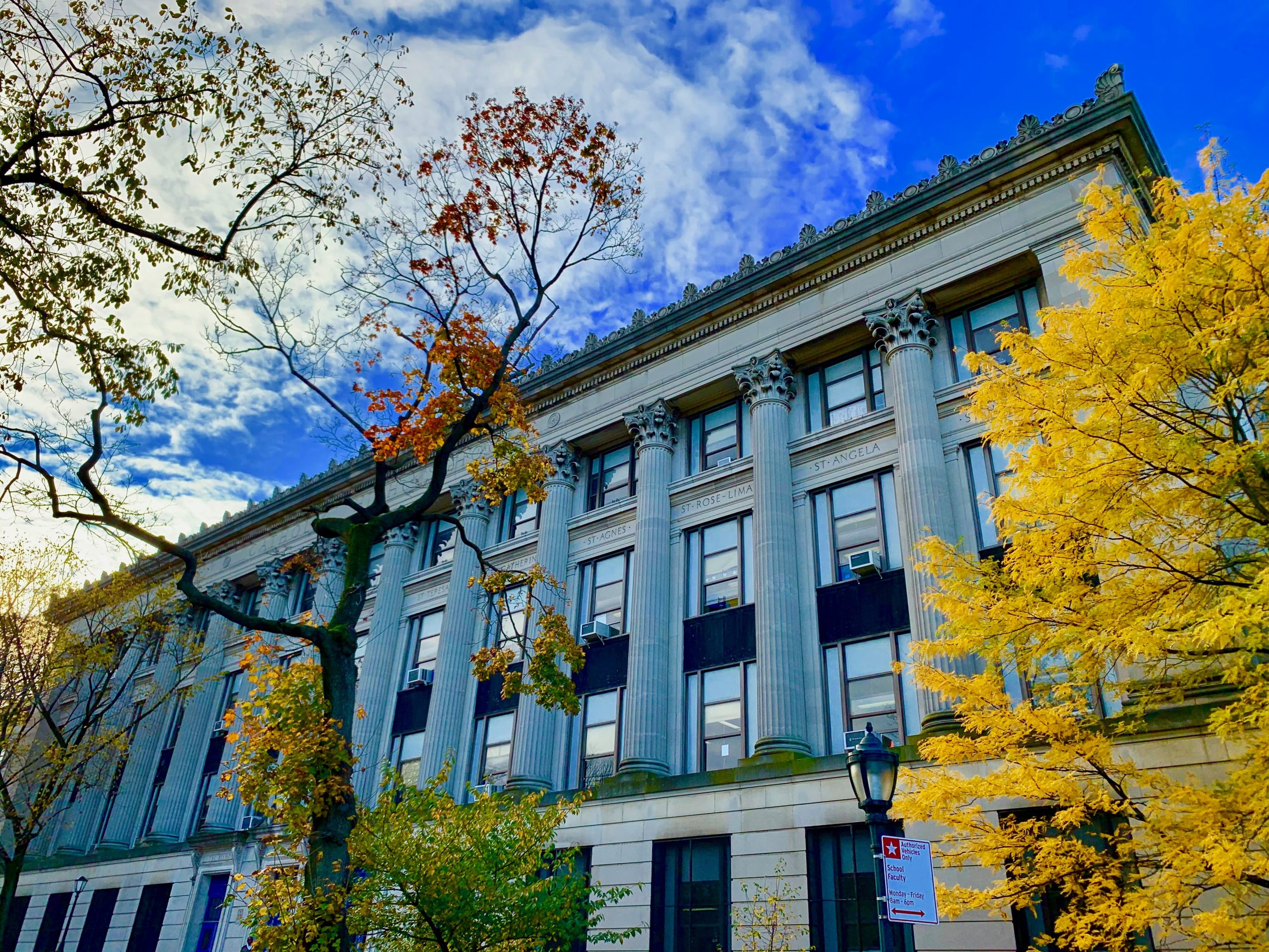 A view of the front of the school with two trees on a Fall day is shown. One tree has lost most leaves while the other trees’ leaves are all yellow. The angle of the shot is looking up as we see many pillars of the building’s architectural style and a partly cloudy blue sky.
