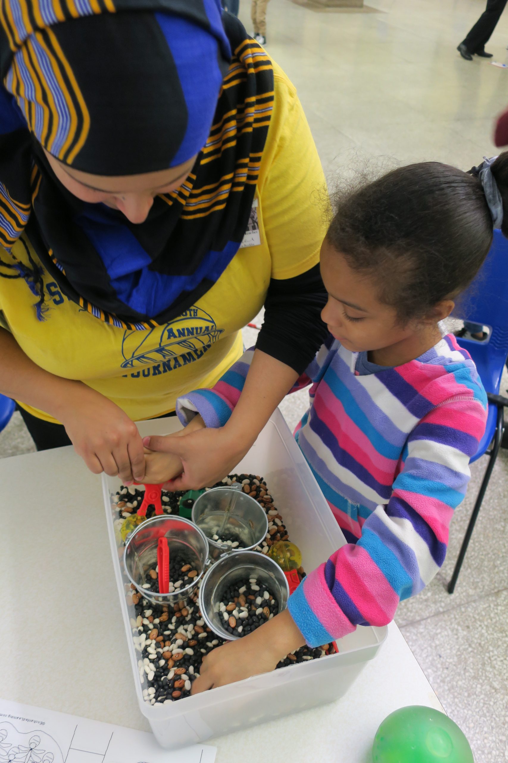 A female teacher and a female elementary student are working on an activity with their hands in a bin with beans. The teacher uses hand over hand to support the student on how to use the scooper to move the beans from the large pin to the small cups.