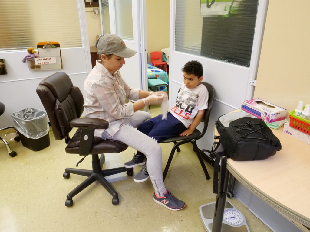 A female school nurse is sitting on a chair facing beside a male elementary student in the nurse’s office. The student is watching the nurse put on disposable gloves.