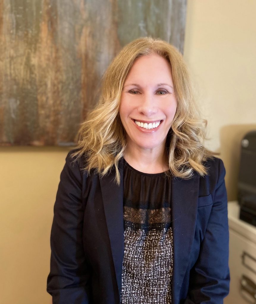 Jodi Falk, Executive Director, is sitting in an office and smiling at the camera. She wears a navy blazer and has blonde hair. There is modern art decor on the cream wall and a printer on the top of white file cabinet in the background.
