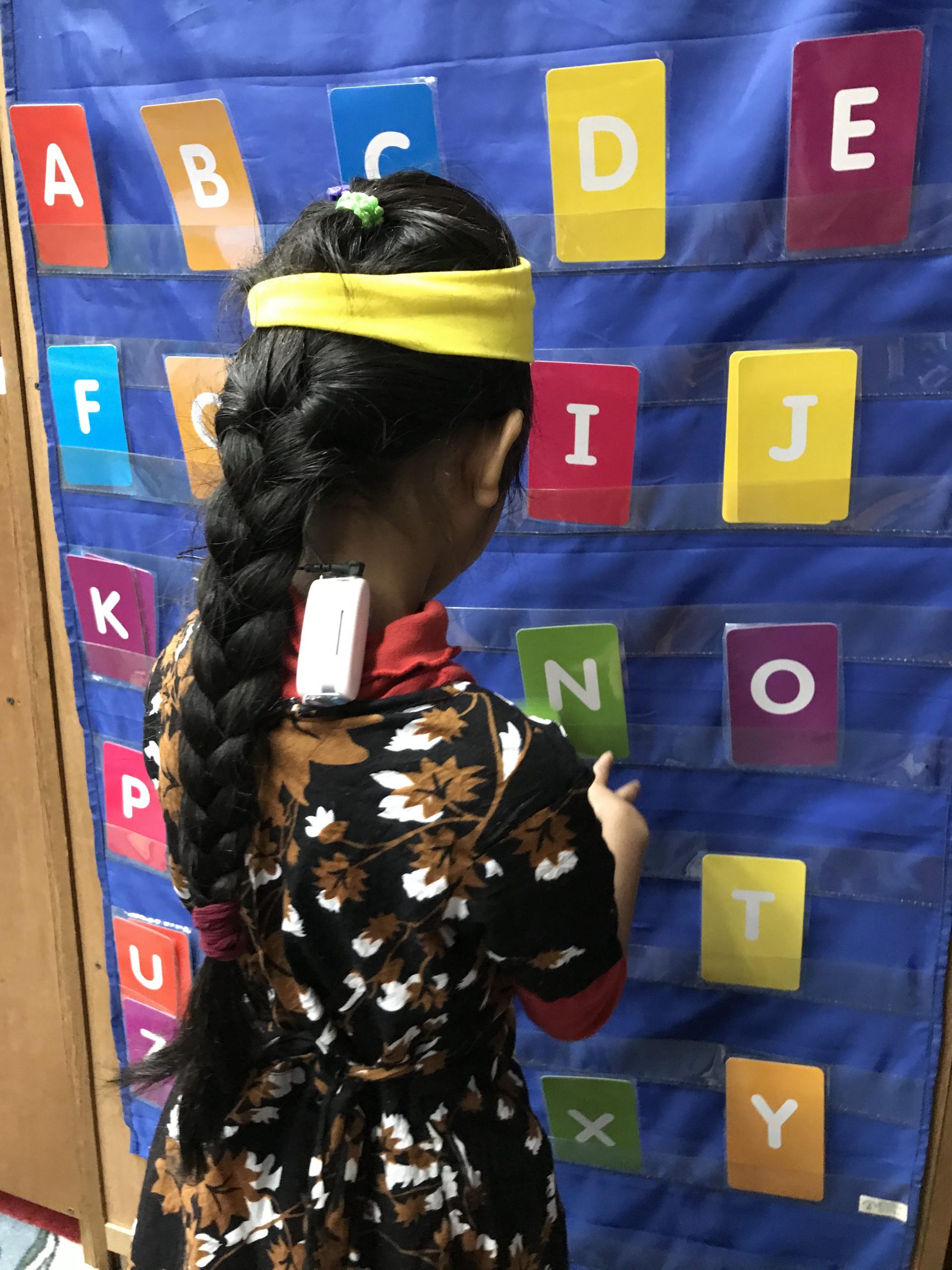 The back of an elementary school female student is shown as she stands in front of a blue pocket chart containing uppercase letters of the alphabet. She is wearing a dress and her long brown hair is in a braid. She wears a yellow headband and her speech processor is clipped to her shirt.