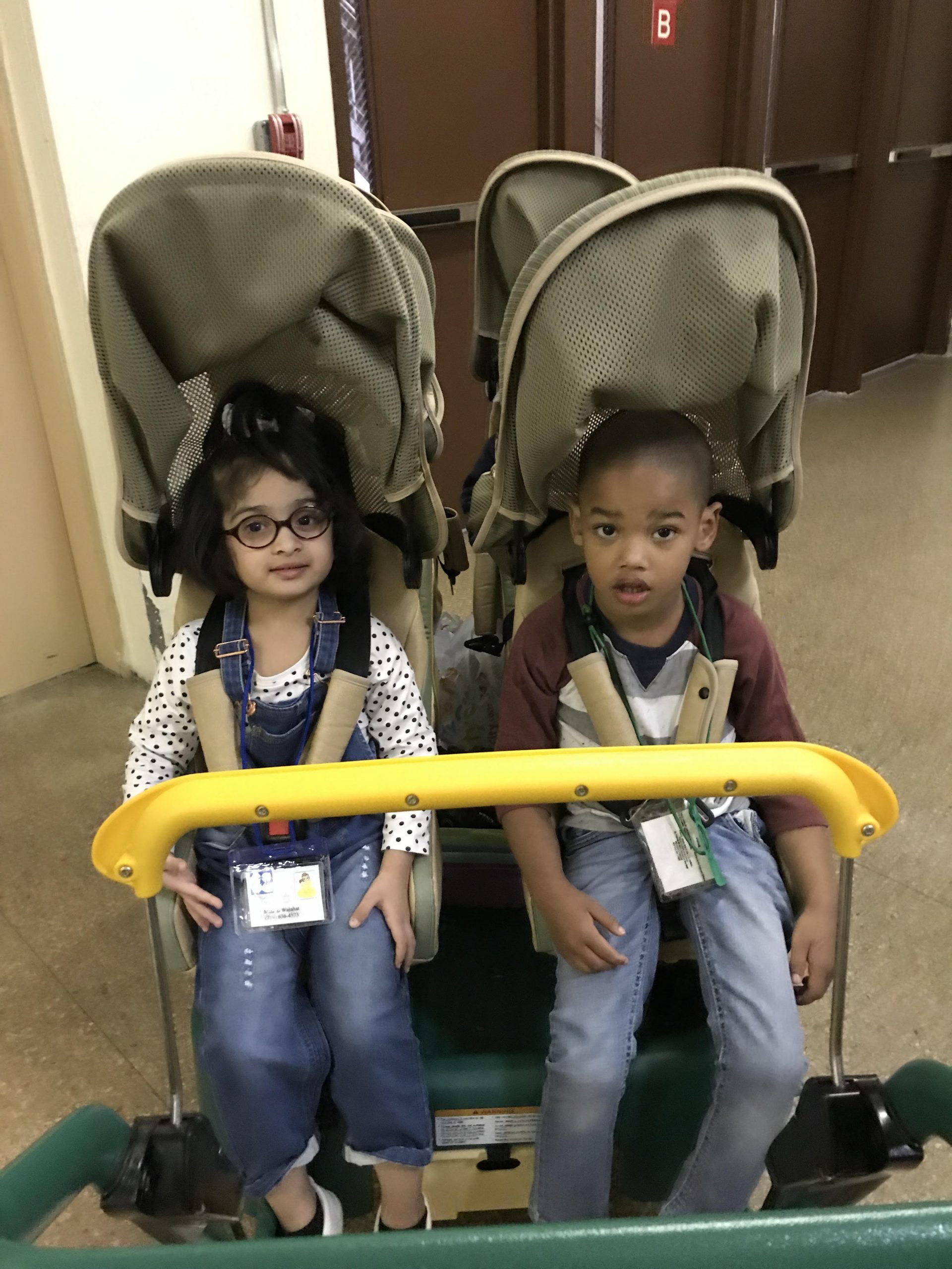 Two preschool students (1 boy and 1 girl) are sitting buckled into a 4 -person buggy stroller hallway right by exit doors. The students are wearing their school IDs around their necks. The female student is wearing eyeglasses, blue denim overalls, top with long sleeves and black dots. The male student is wearing a top with long sleeve style sports sweatshirt and bottom denim jeans.