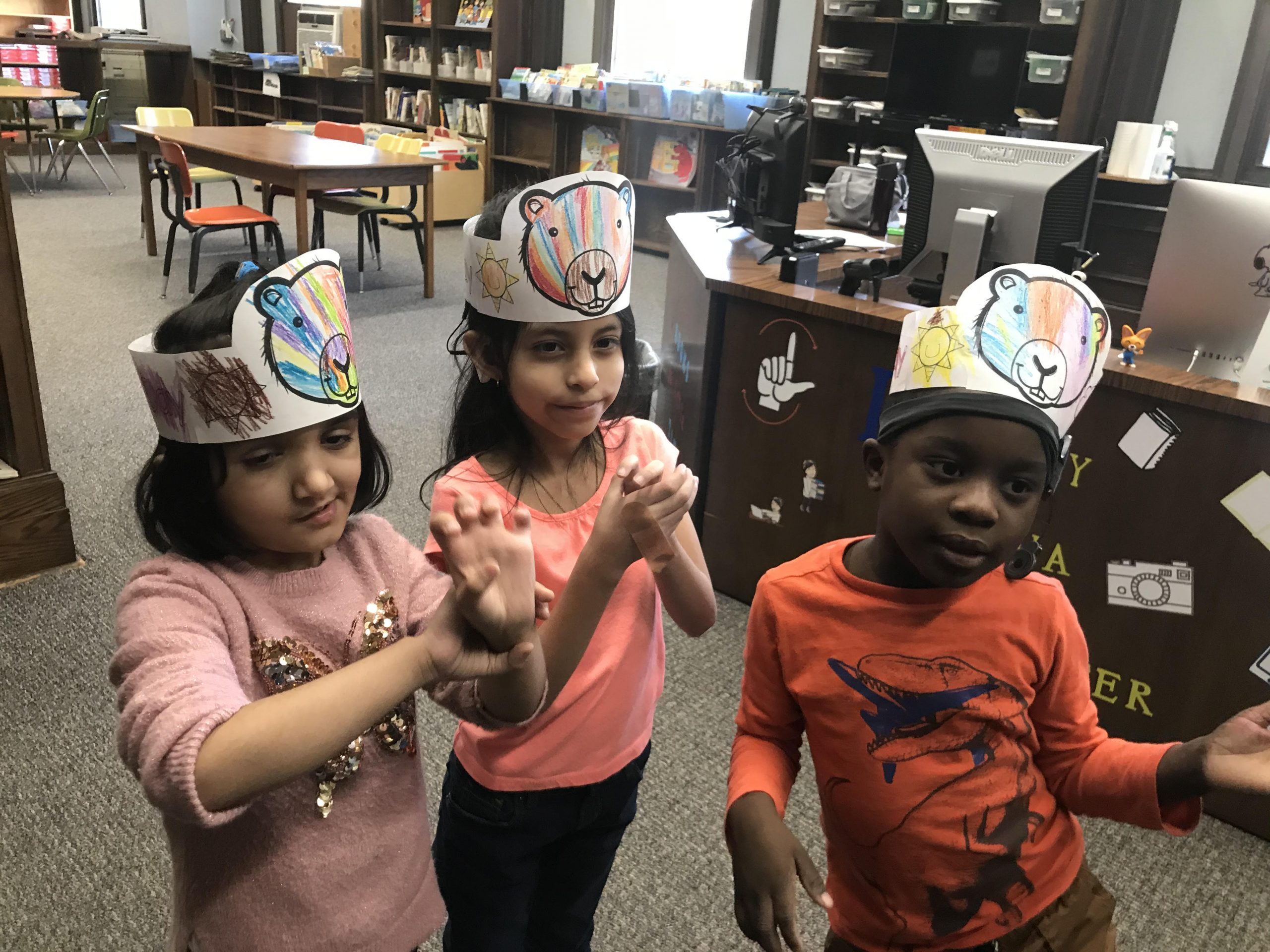 Three preschool students (2 girls and 1 boy) wearing Groundhogs Day paper crowns on their heads are in the library. They are all signing at the same time.