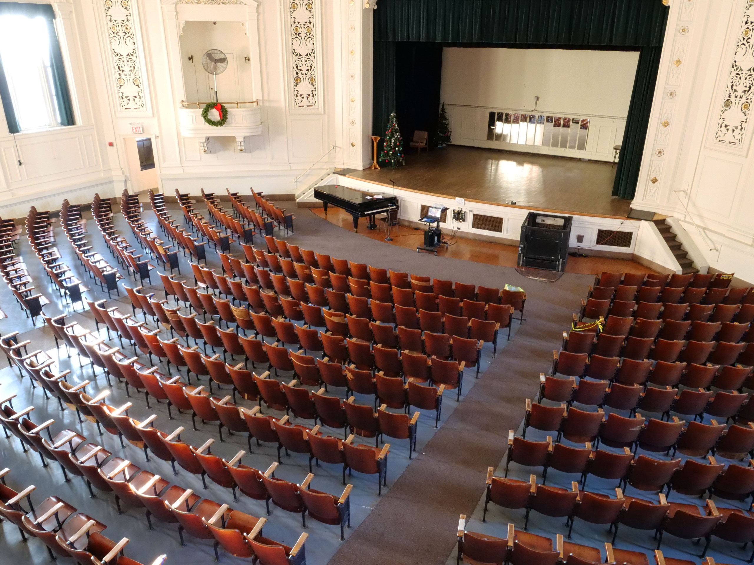 In an aerial view of the school auditorium we look down to view many wood folding seats, two small balcony boxes, Christmas wreath hanging on the balcony box, black concert grand piano, laptop and project equipment, microphone, vertical wheelchair lift, theater stage, wooden lecture stand, two Christmas tree stands on an auditorium stage and rectangular wall mirrors back of the stage in the background.