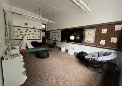 A small multisensory room with brown carpeting, yoga mats, beanbags, large circular chair & small cubbies with books. The bulletin boards are decorated with posters.