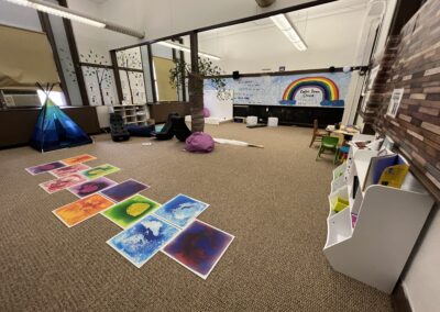 View of a classroom with carpeting. In the center there is a pole wrapped & decorated as a tree. There are gel pads, beanbags, rocking chairs, yoga mats and a tent. On the back wall is a bubble mirror. On the side wall there are cubbies and a small table & chairs.