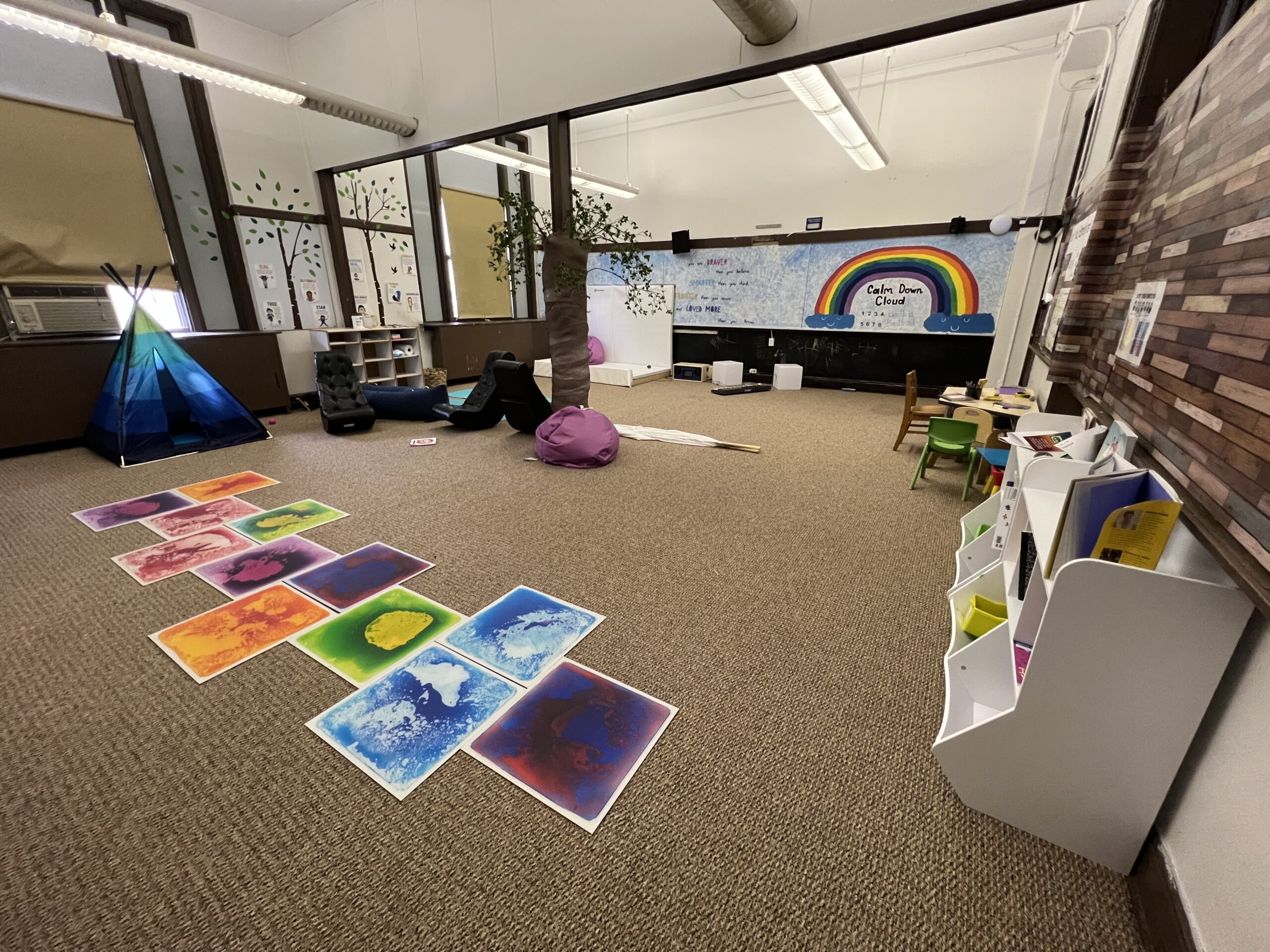 View of a classroom with carpeting. In the center there is a pole wrapped & decorated as a tree. There are gel pads, beanbags, rocking chairs, yoga mats and a tent. On the back wall is a bubble mirror. On the side wall there are cubbies and a small table & chairs.