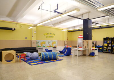On a cafeteria wall, a sign is painted Literacy Lounge. There is a long couch, rugs, flexible seating, tents, beanbags, desks, and a writing center topped with baskets of paper and supplies.