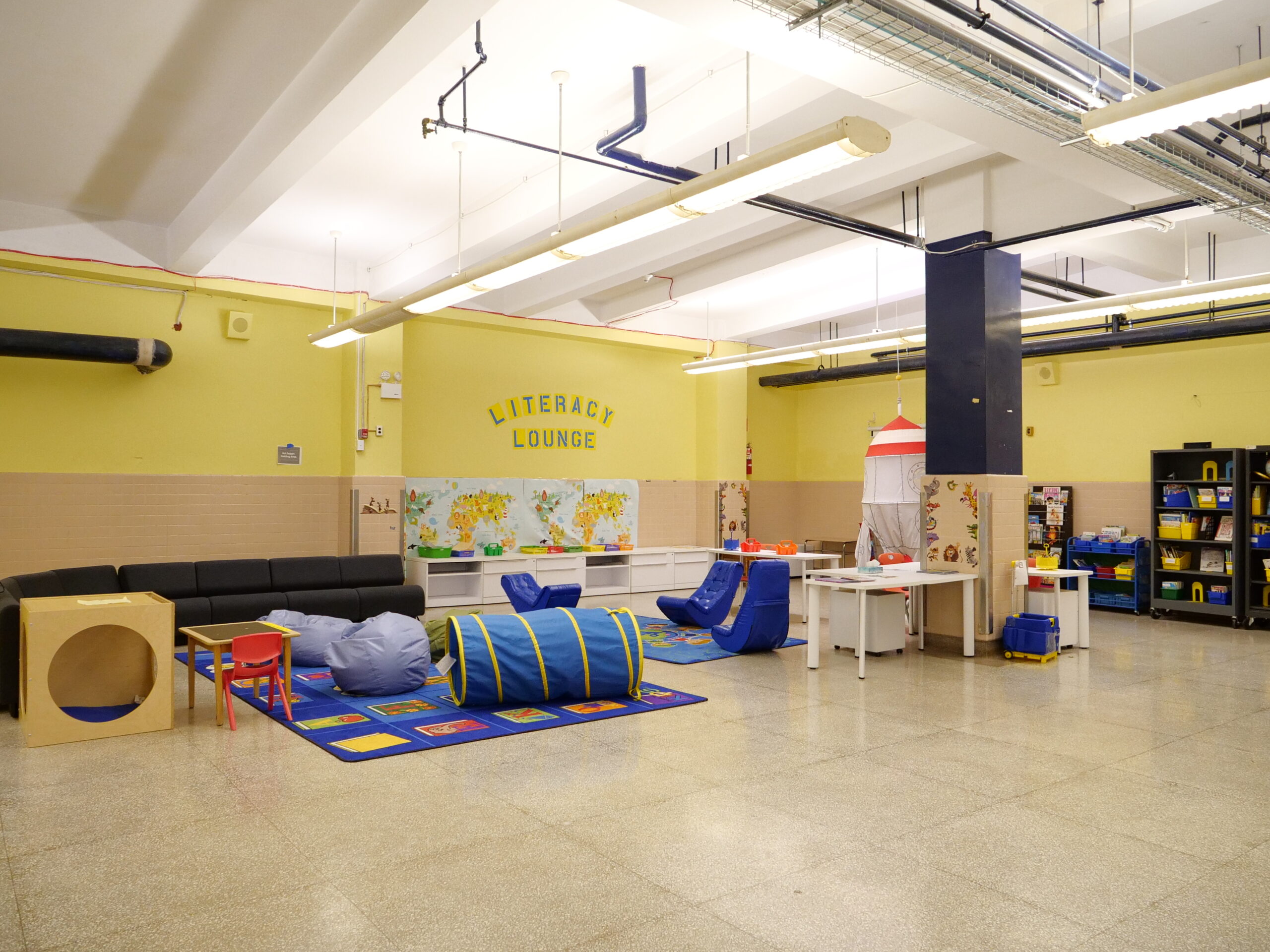 On a cafeteria wall, a sign is painted Literacy Lounge. There is a long couch, rugs, flexible seating, tents, beanbags, desks, and a writing center topped with baskets of paper and supplies.