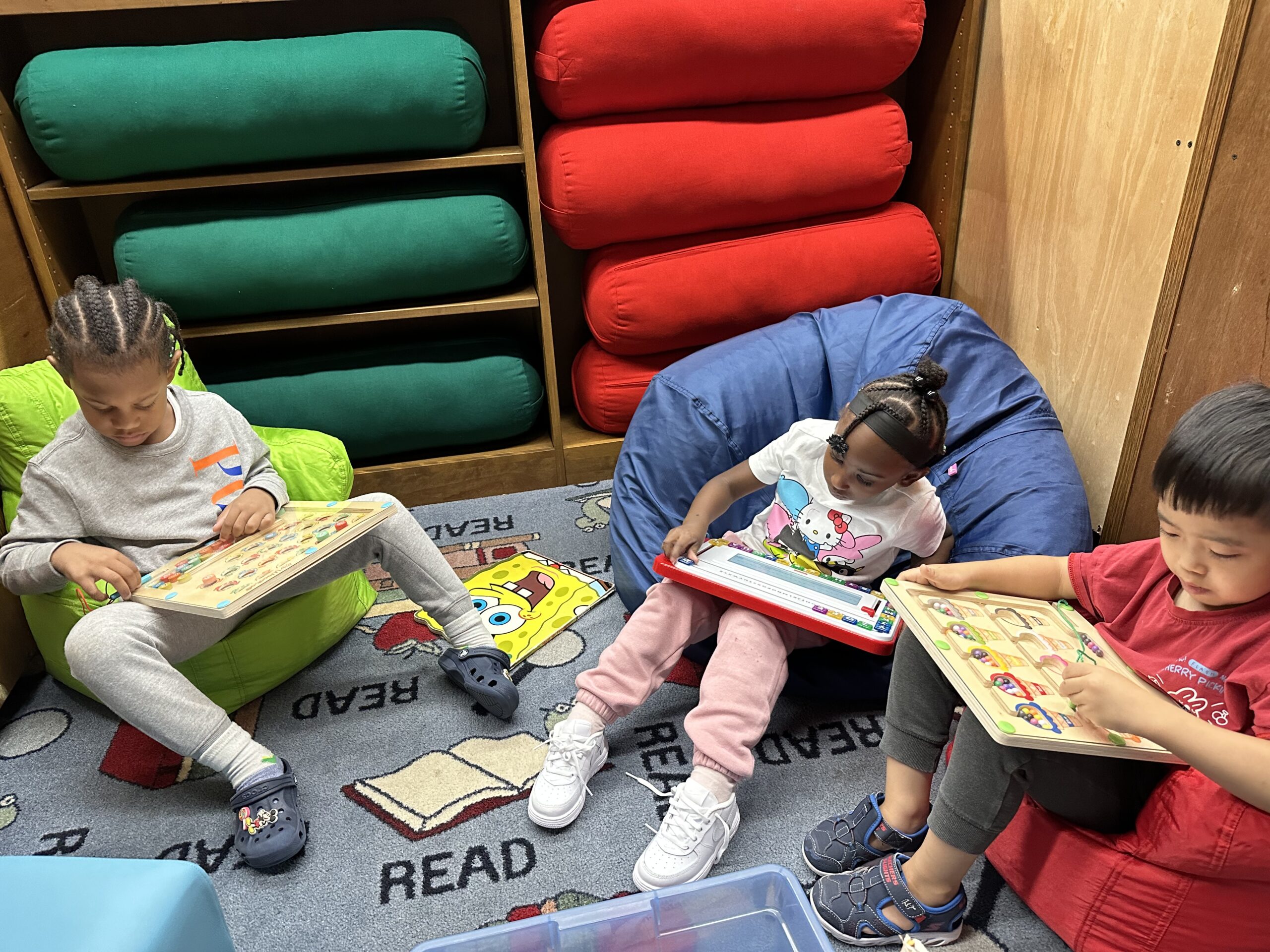ID: 3 Preschool students sit on bean bag chairs in a reading nook solving puzzles independently.