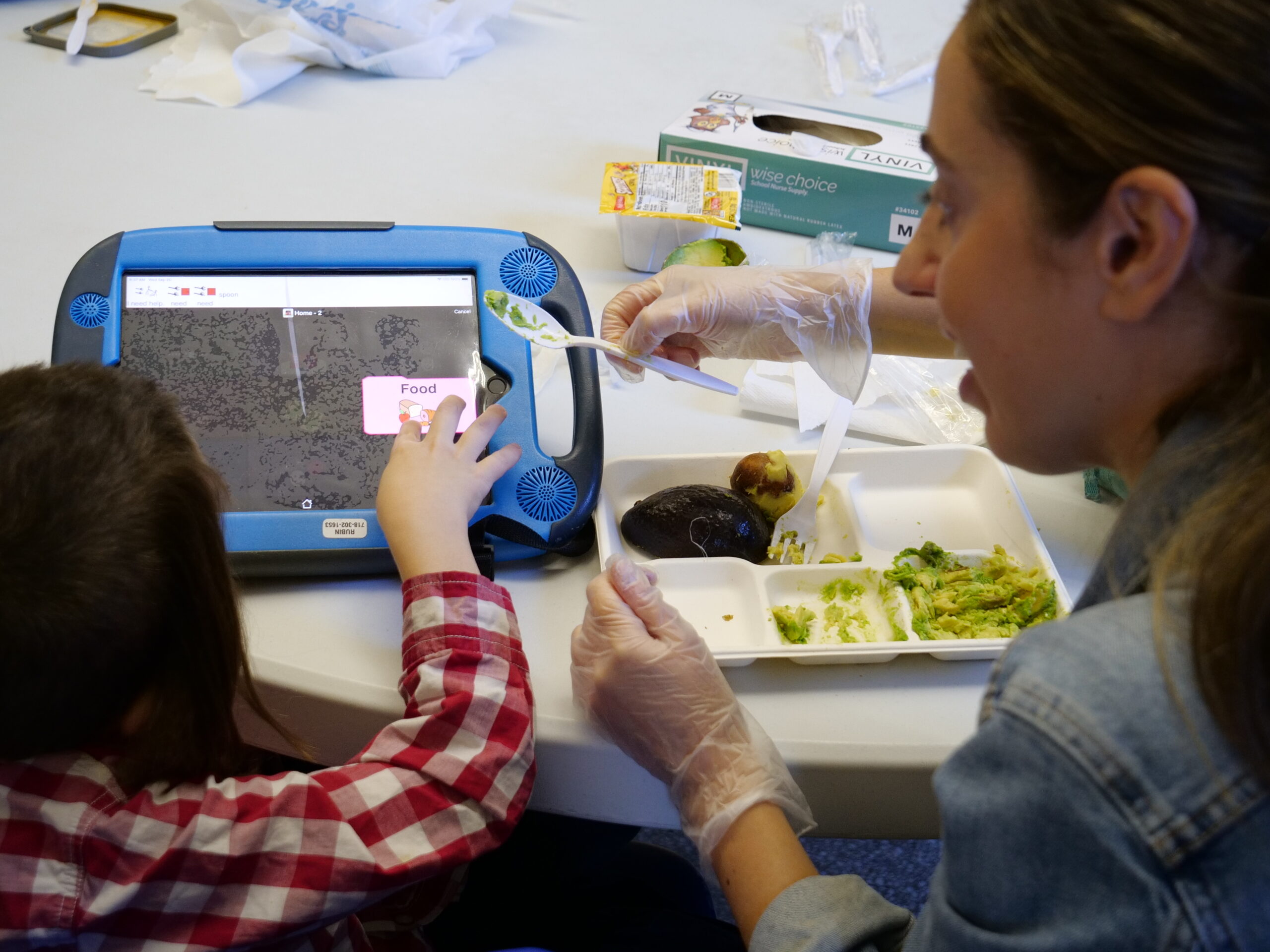 ID: A student points to the word and image of "food" on his AAC device. His feeding therapist sits next to him with a tray of mashed avocado while holding a spoon.