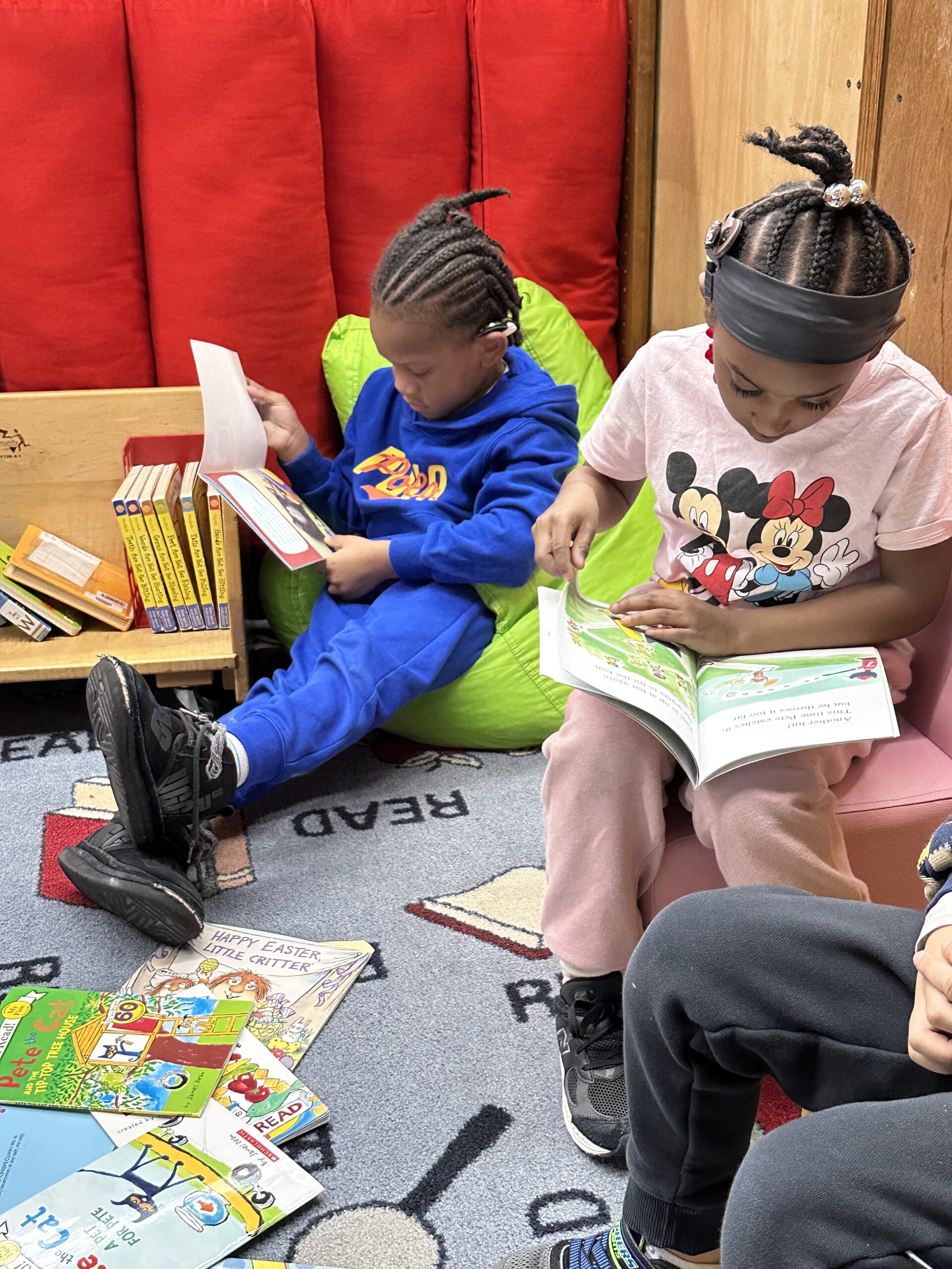 Two young students sit in the reading nook of a classroom each reading a book independently.