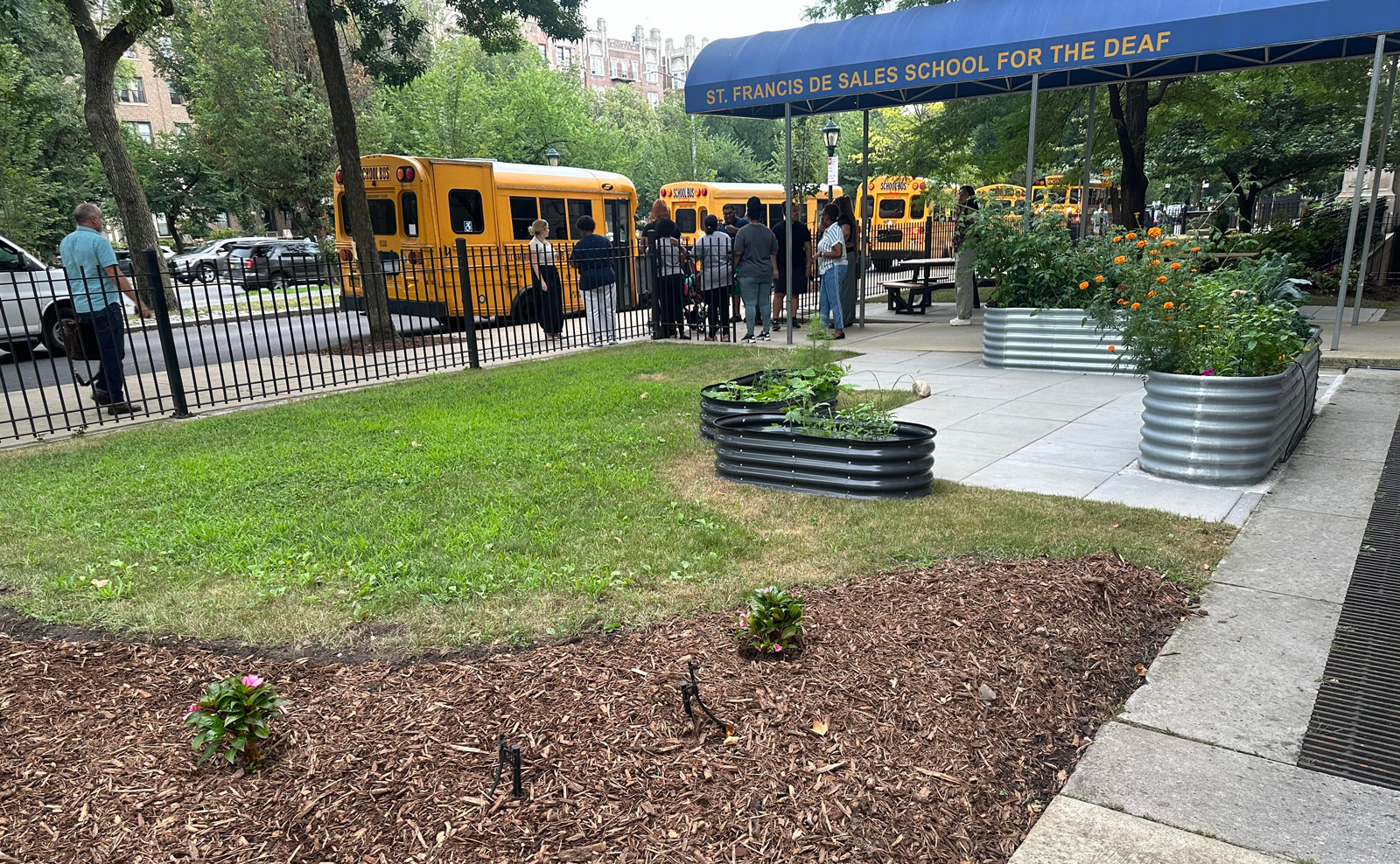 Staff standing outside the Accessible Entrance with a long blue awning. The school garden is in the forefront of the picture.