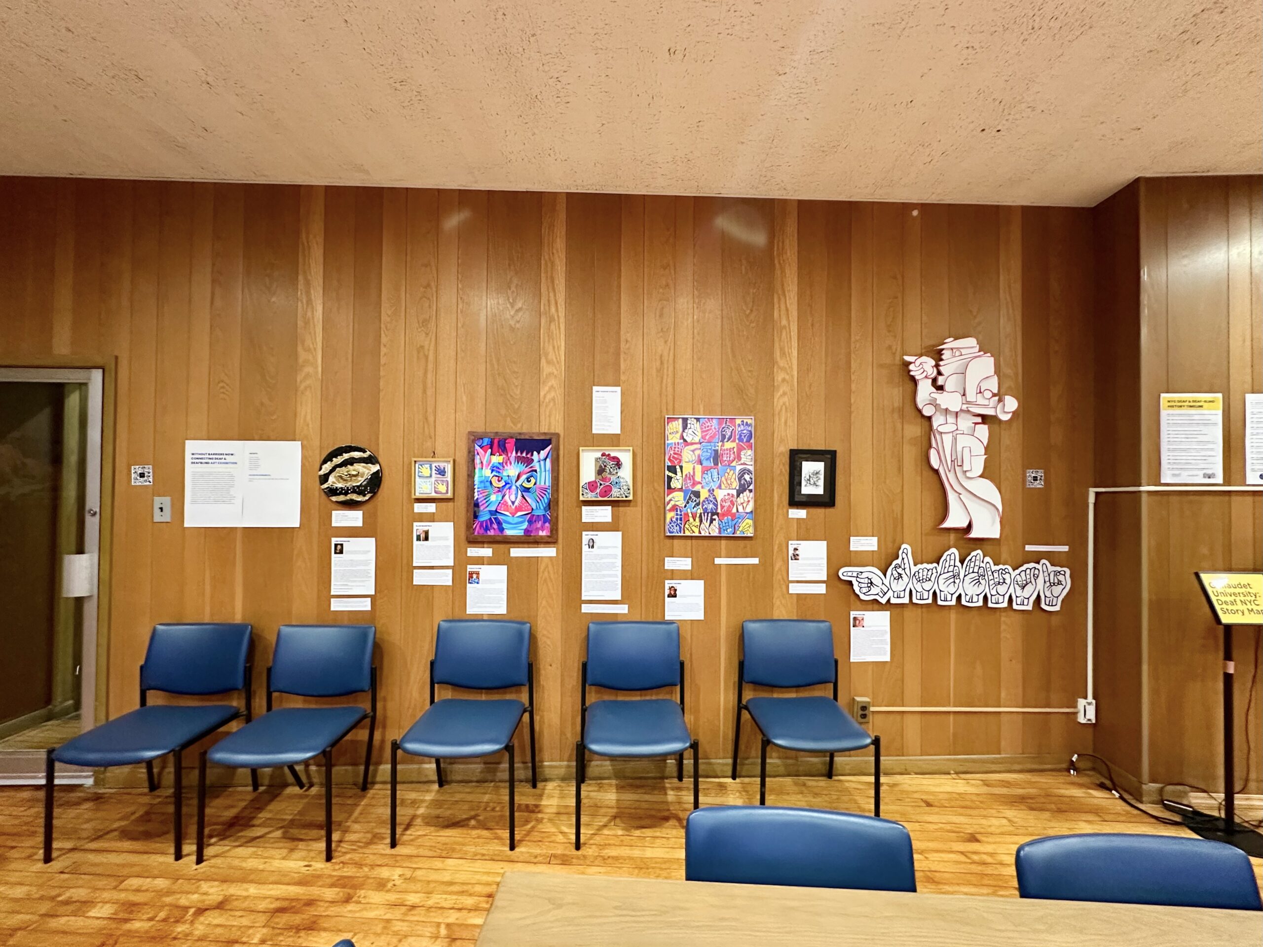 Museum wall display of documents and artwork with a row of chairs set out in front for visitors.