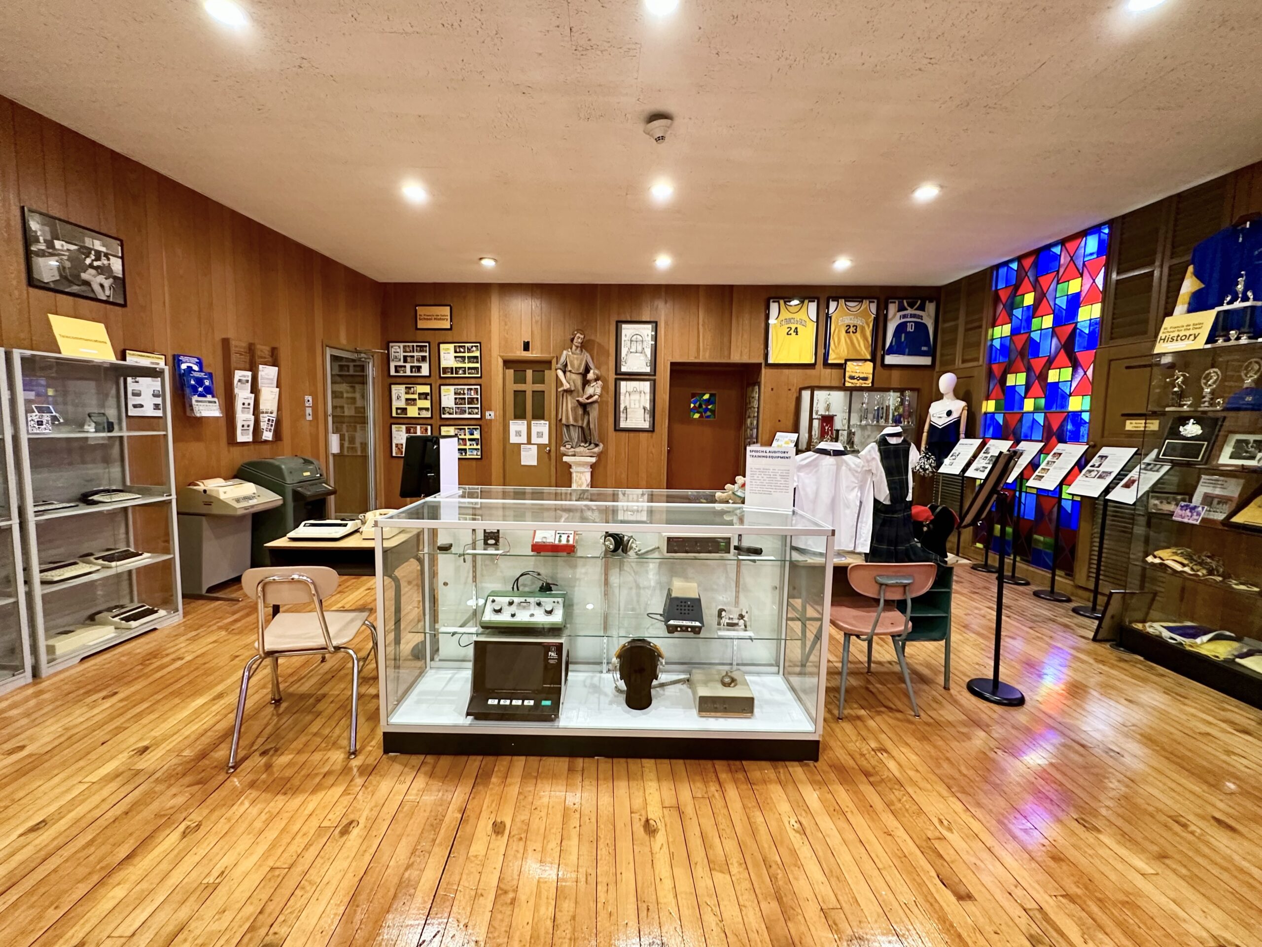 Museum gallery with glass display cases and a wall of coloured stained glass panels.