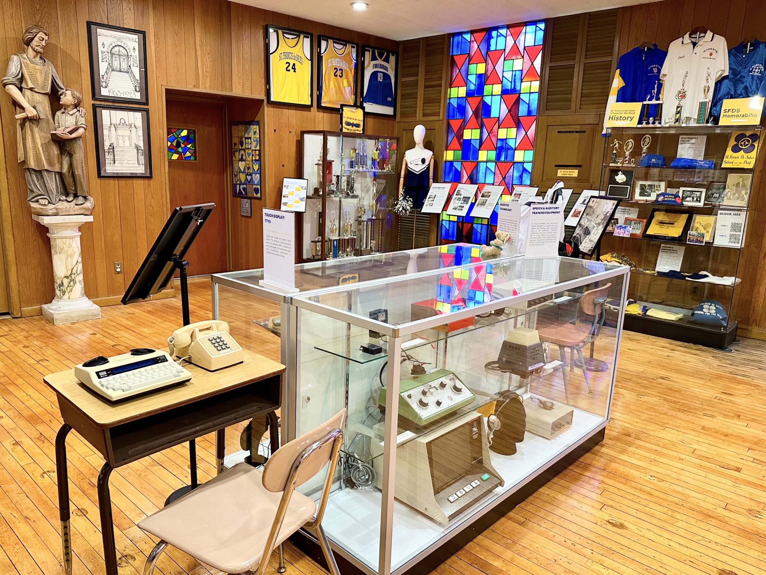 Museum exhibit with a glass case of objects in the foreground and a religious statue and artwork on display behind it.