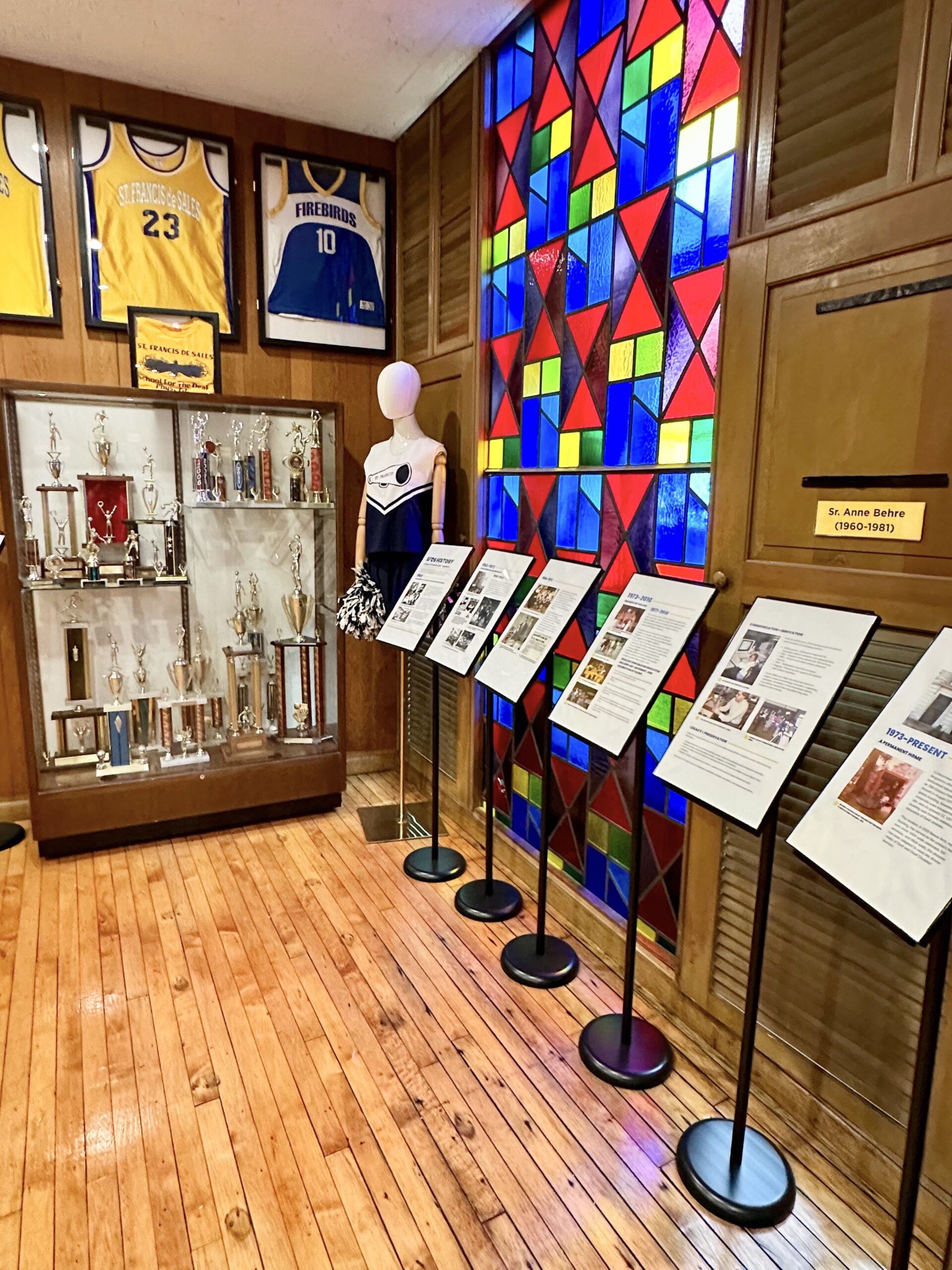Museum wall of coloured stained glass panels above a row of framed documents and display stands.