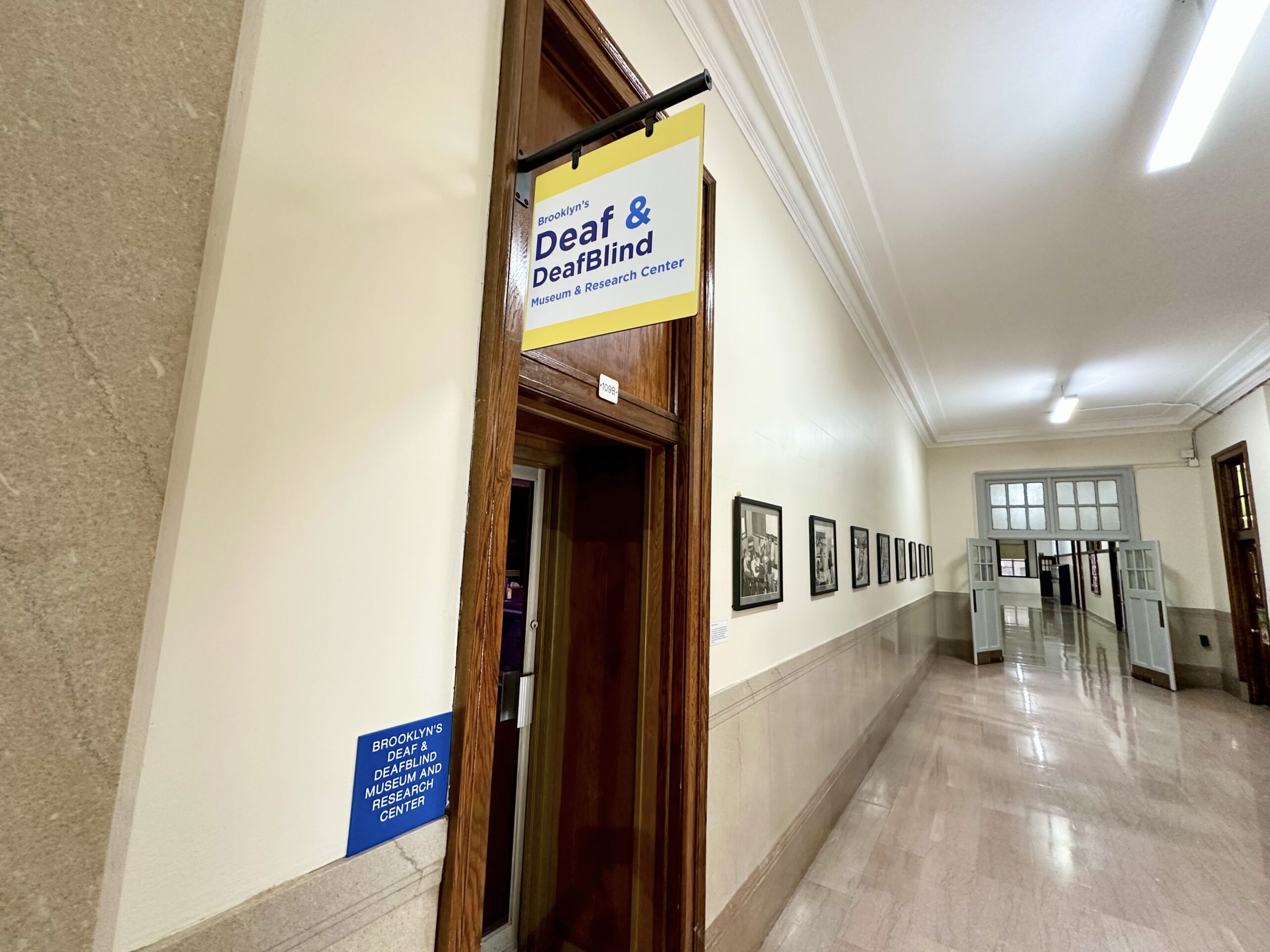 Corridor leading to the museum, with a yellow Deaf and DeafBlind sign above the doorway and framed photographs along the wall.