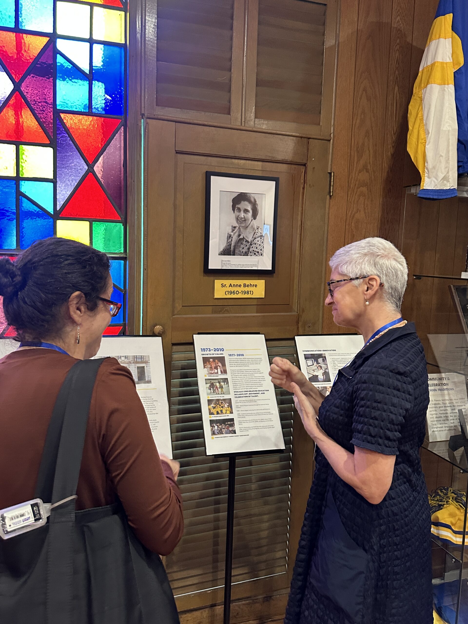 Two visitors reading the framed exhibits in the museum beside a wall of coloured stained glass.