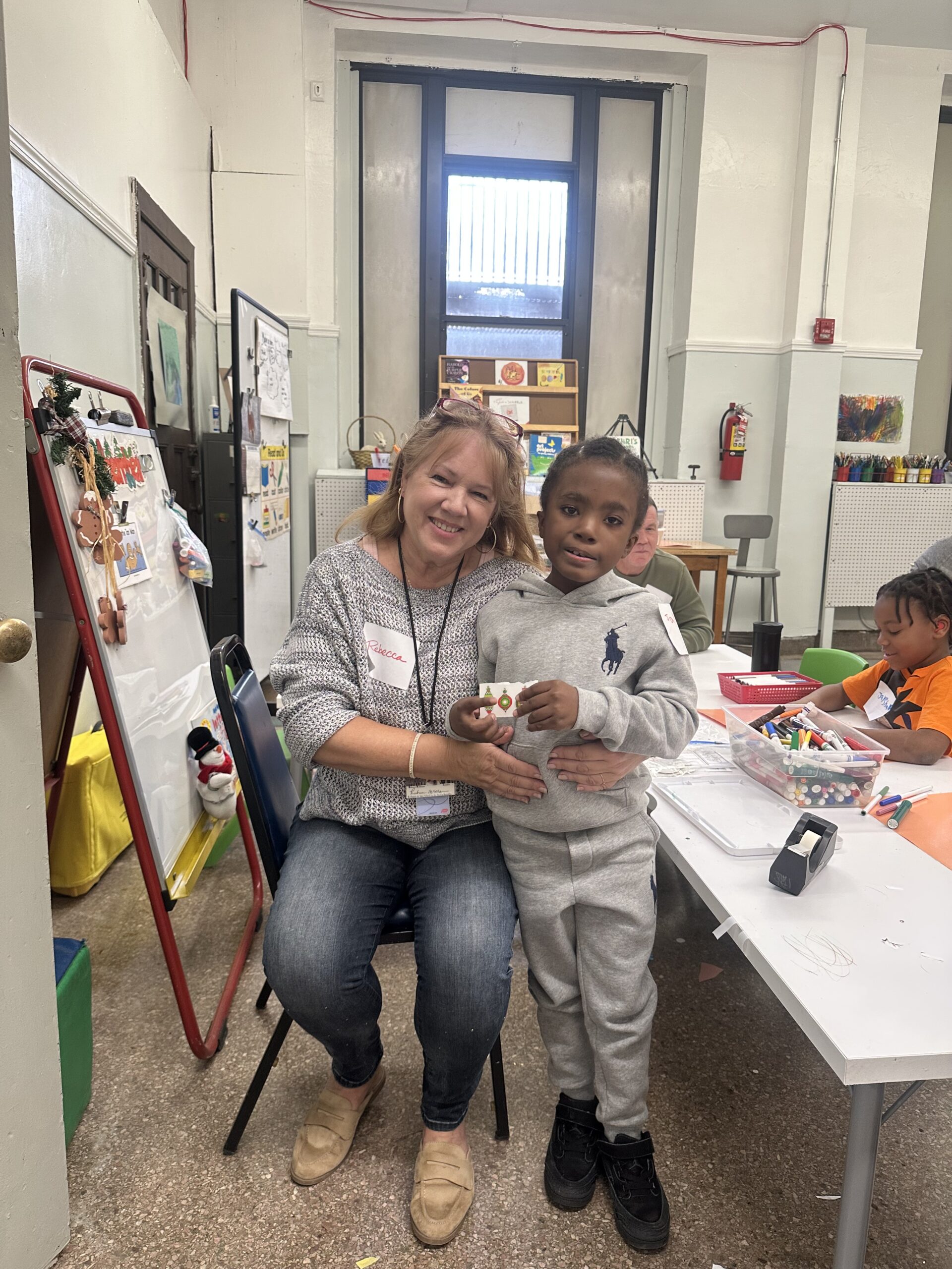 A volunteer and a young student smile together at the art table, holding a handmade craft.