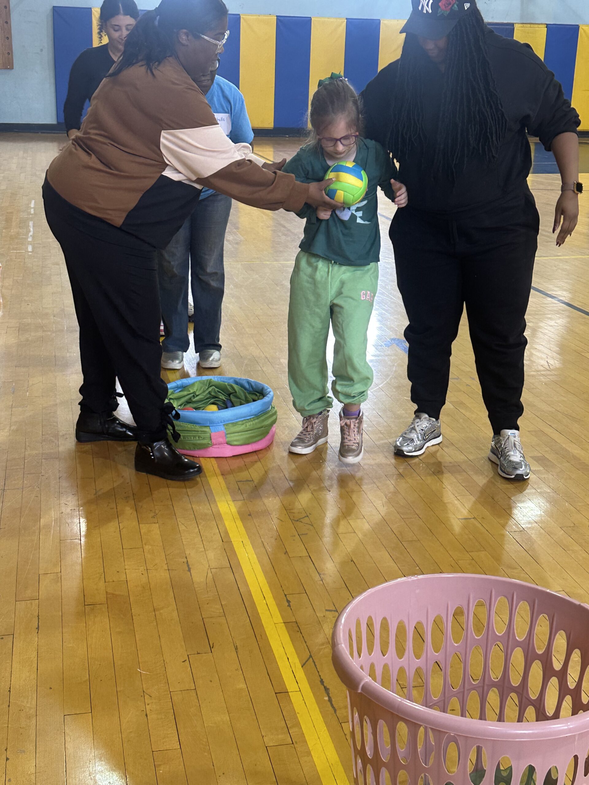 A student holds a ball with support from two staff members during an adaptive sports game in the gym.
