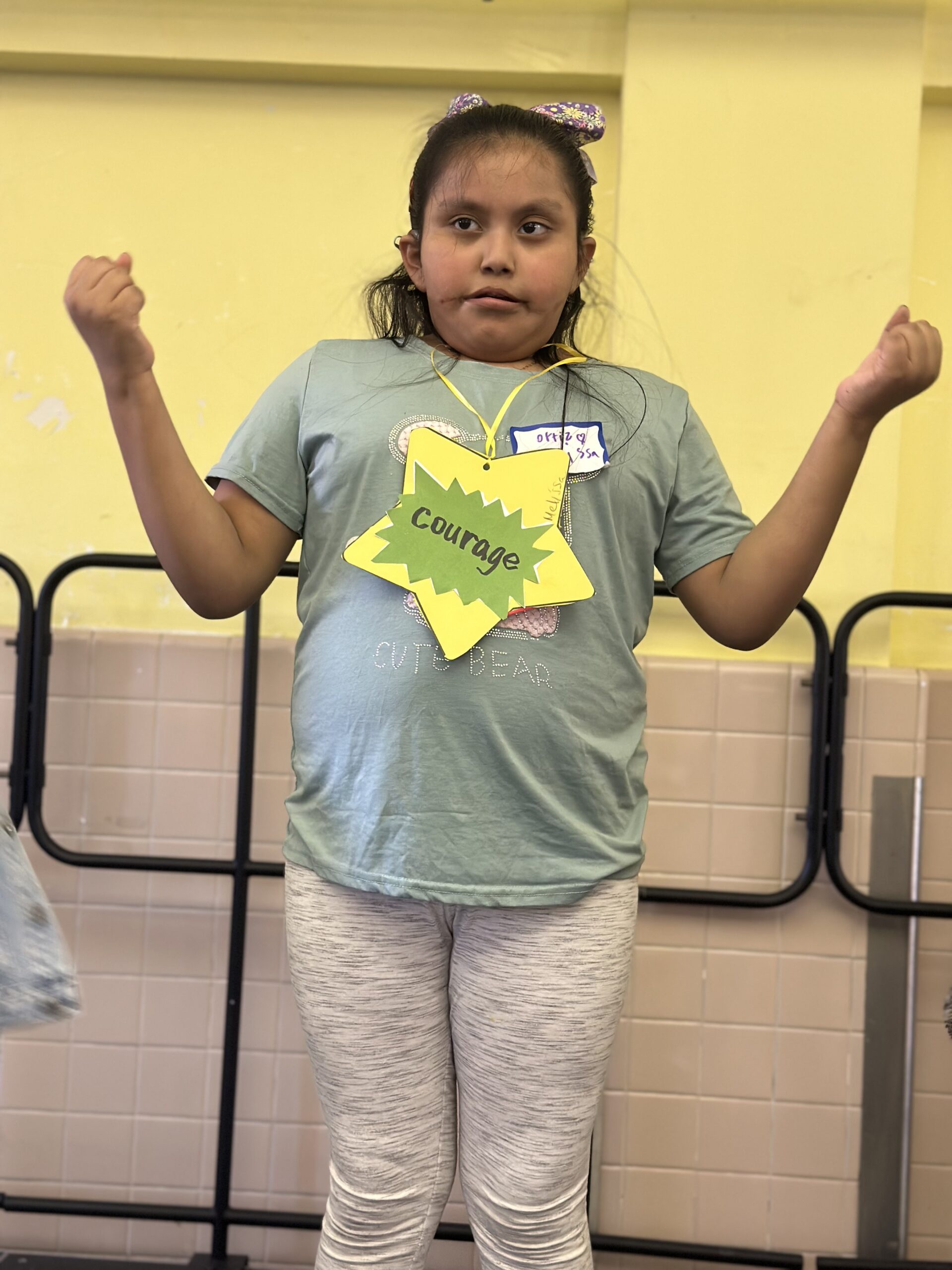 A student wearing a paper star that reads Courage raises both arms with a proud smile.