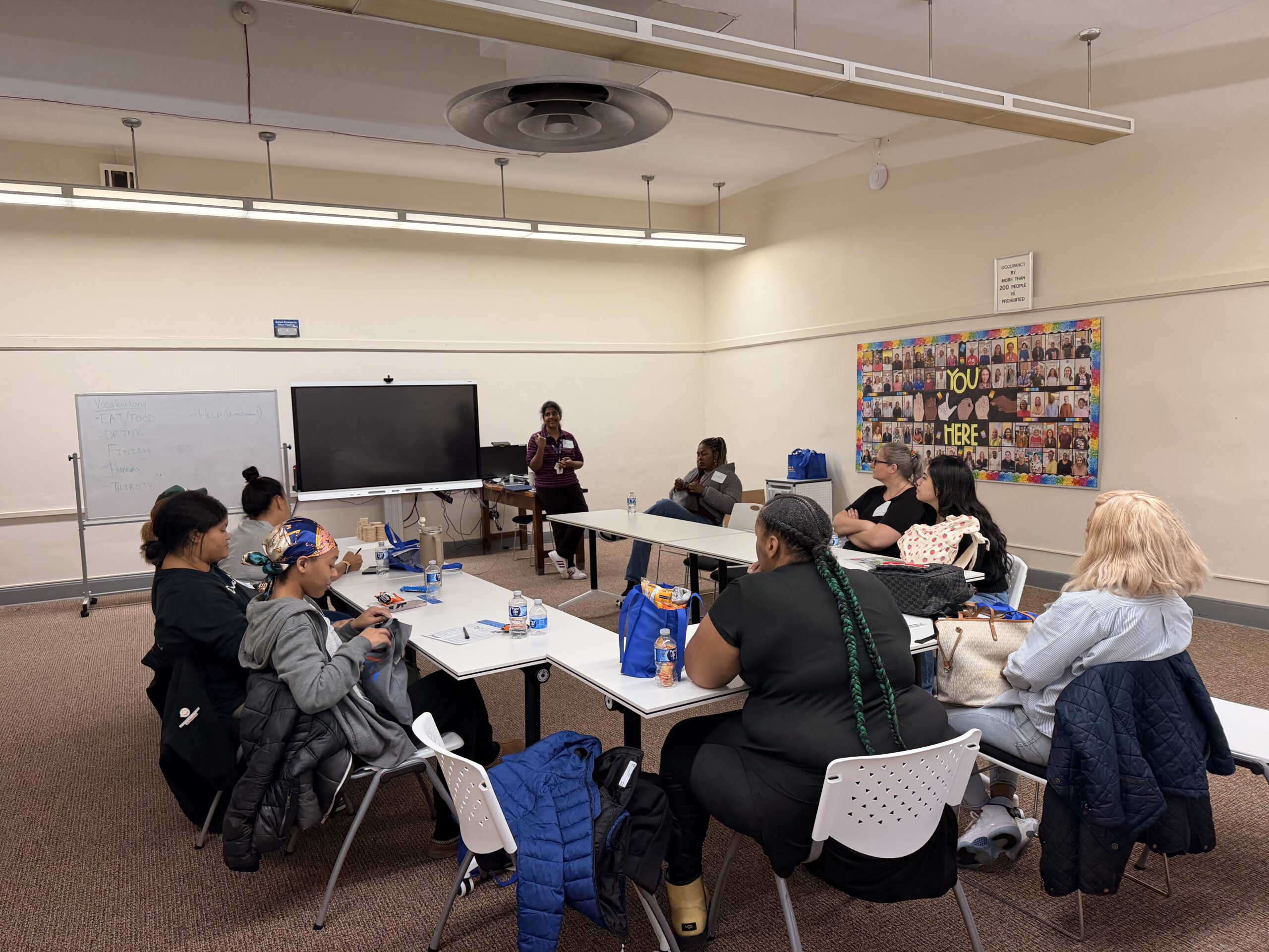 Parents and caregivers gather around tables for an ASL vocabulary lesson while a Deaf educator signs at the front of the room.