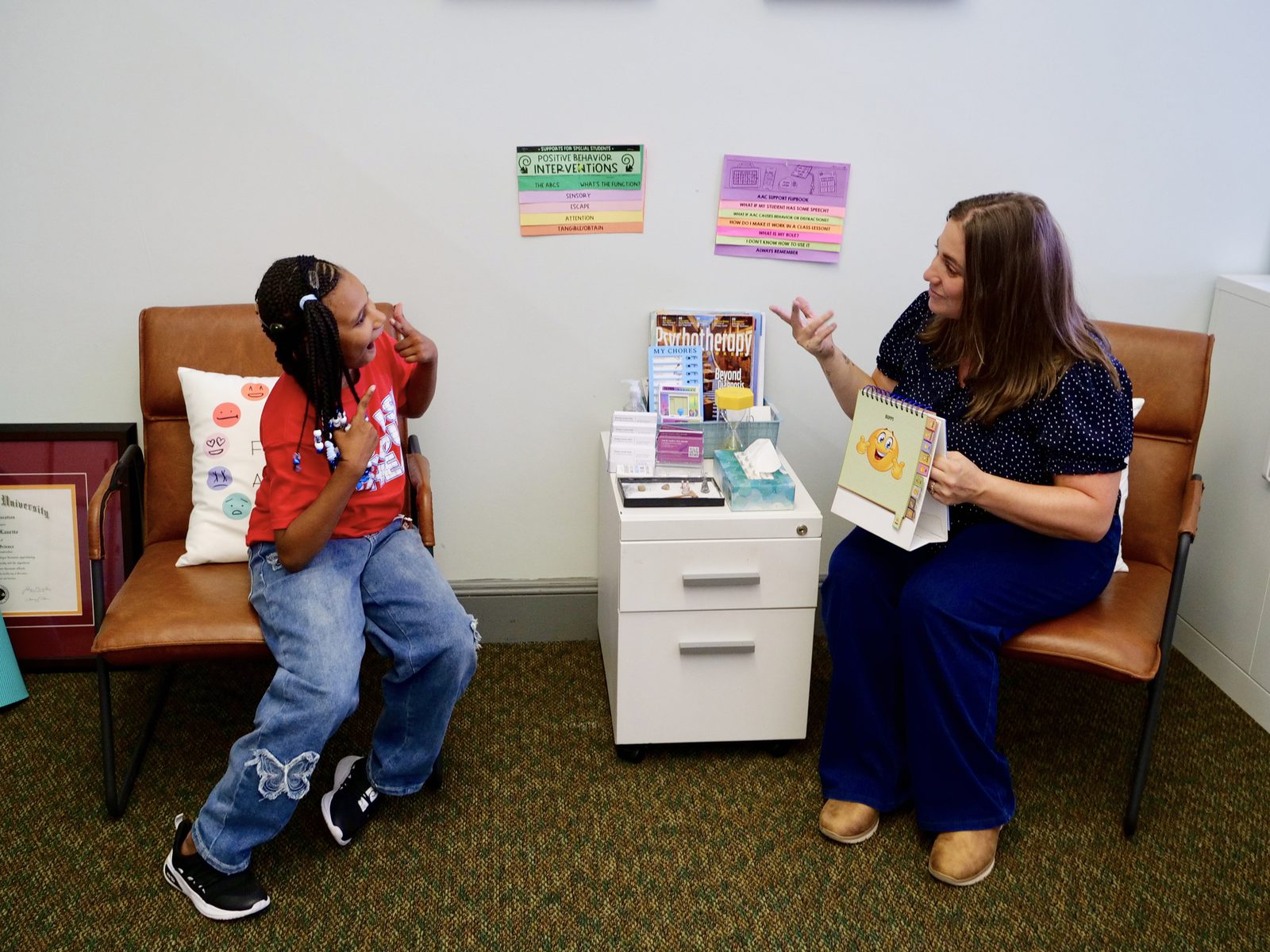 A school counselor and a student sit facing each other in armchairs, both signing, with books and materials on a cabinet between them.