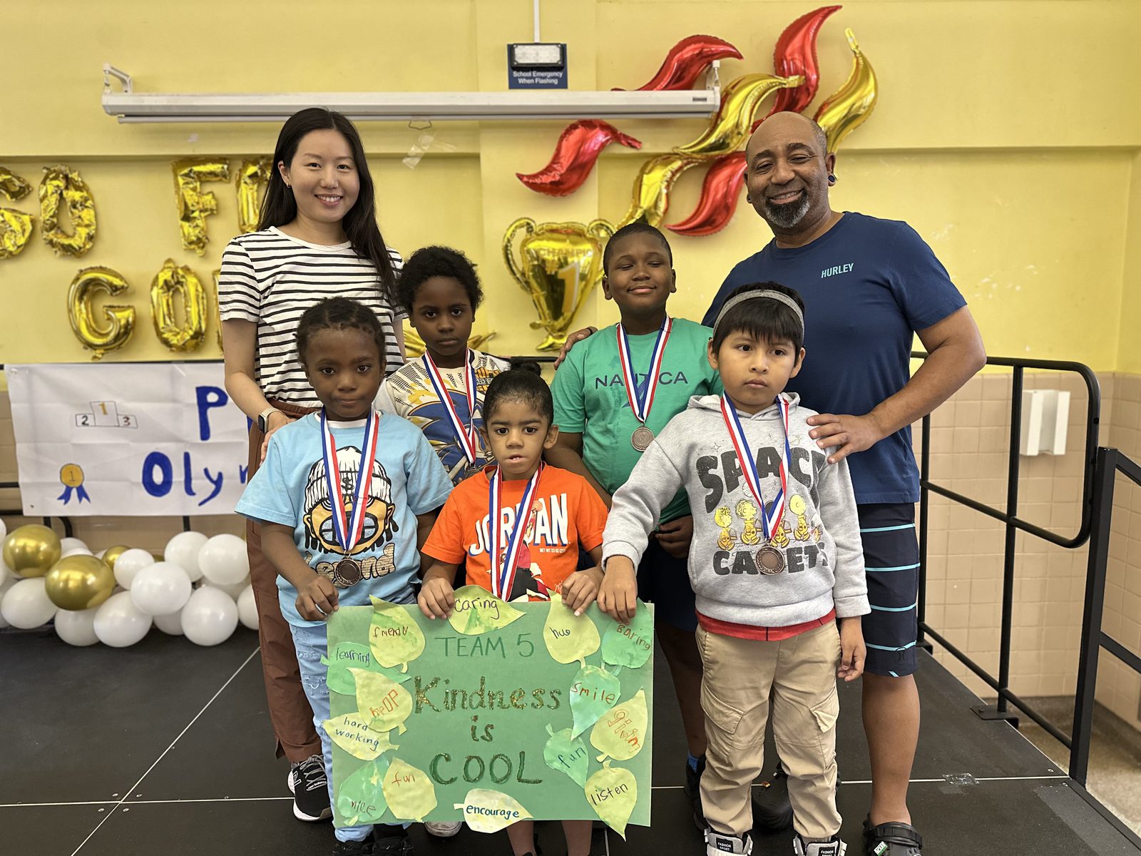 Five students wearing medals hold a poster reading TEAM 5, Kindness is COOL, with two staff members standing behind them at the PBIS Olympics.