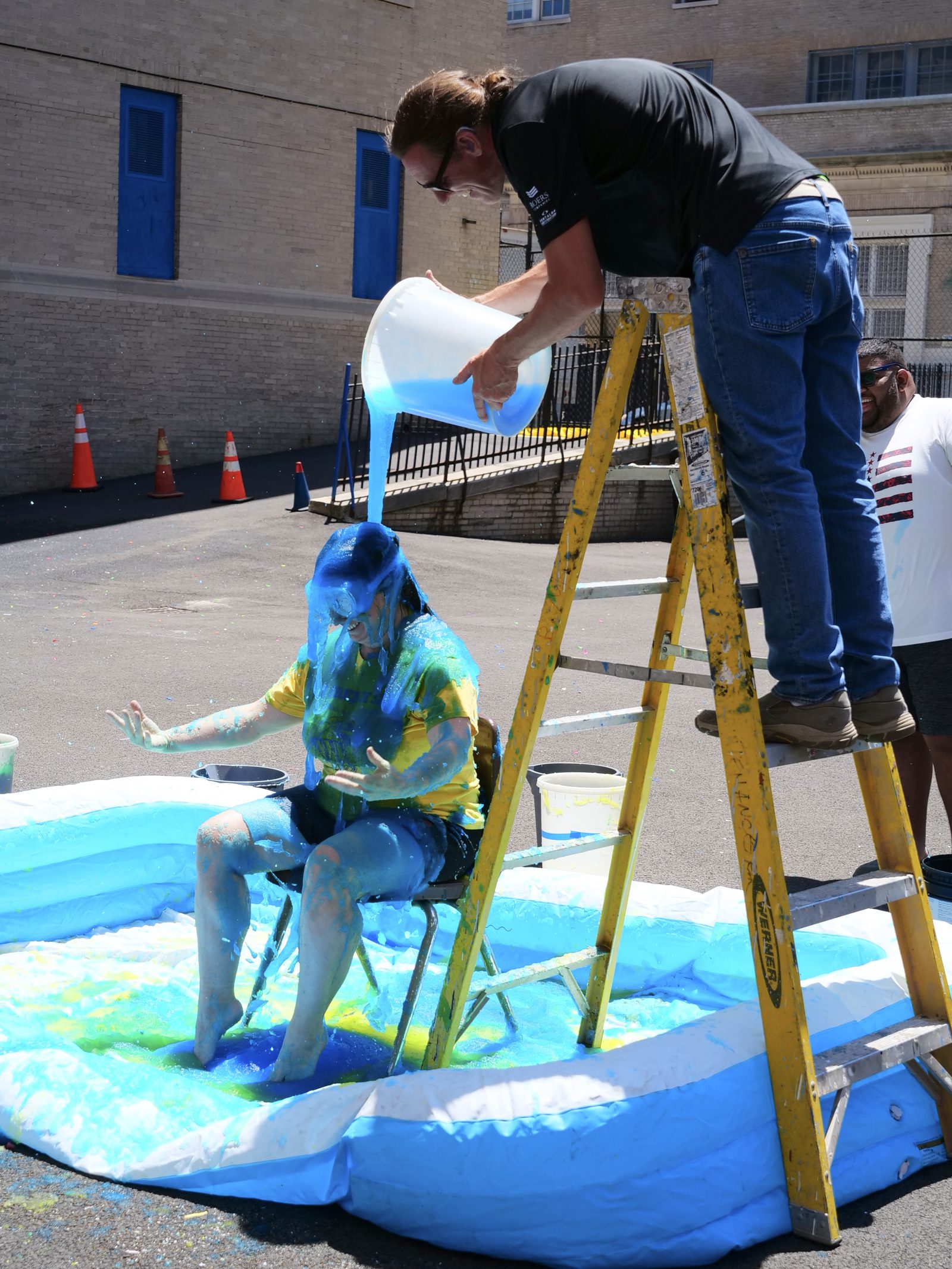 A staff member on a ladder pours blue slime over a colleague seated in a paddling pool during an outdoor PBIS celebration.