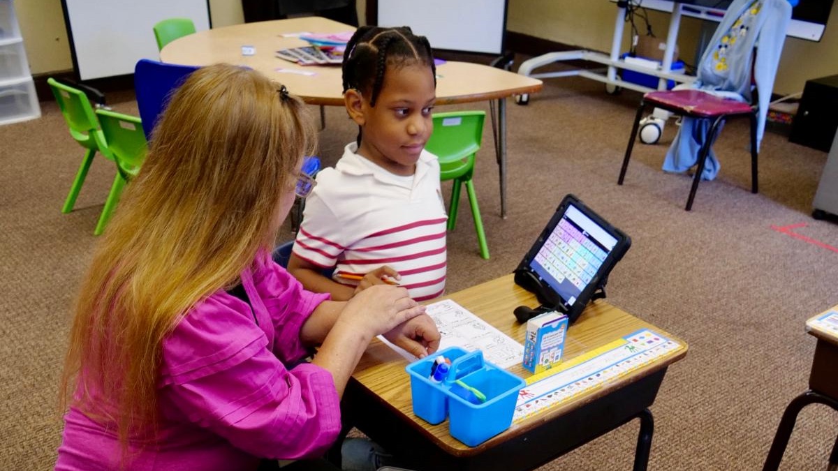 A teacher working one to one with a student who is using a tablet communication device at her desk.